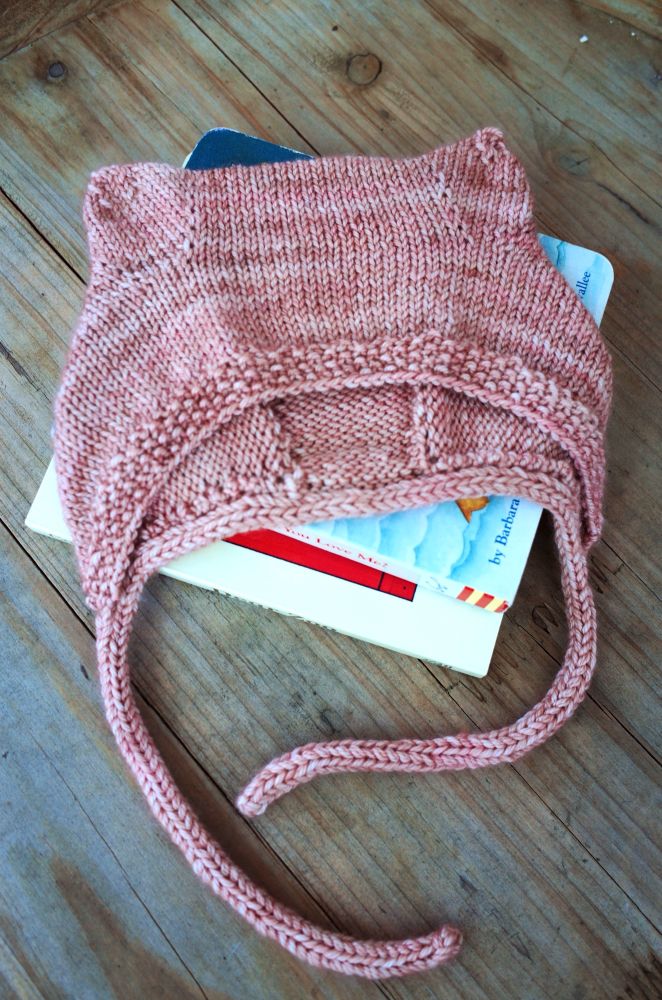 Stockinette baby bonnet with icord ties, moss stitch edging and shaping to make small bear ears at the top. Knit with a pink variegated yarn.