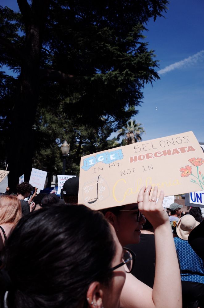A woman shades her face from the son with a sign that reads “ICE belongs in my Horchata not in California”.