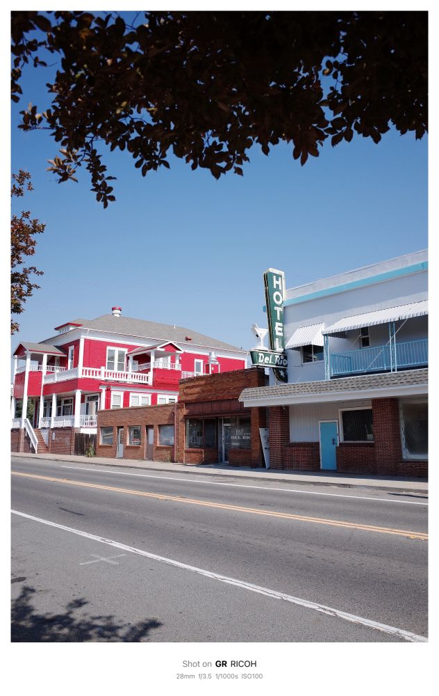 Photo shows an old 1950s era hotel with turquoise trim and a green Hotel sign that reads “HOTEL Del Rio” with a white Martini glass. Next to the hotel is a small brick building followed by a large two story building painted a bright cherry red with white trim on its double porches and gabled roof. The photo is taken from accross the street and the image is framed by tree foliage.

 Shot on GR RICOH
 28mm f/3.5 1/1000s ISO100