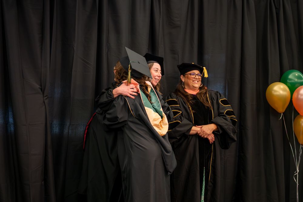 On stage at my MPH graduation, hugging Dr. Aleta Baldwin while Dr. Heather Diaz stands beside us, smiling.