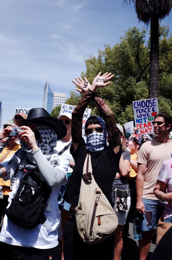 Two women stand in a crowd with their faces covered by Keffiyehs. One holds a small digital camera up to her face. The other looks into the camera with her arms crossed over her head. “FUCK ICE” is written accross her palms. 