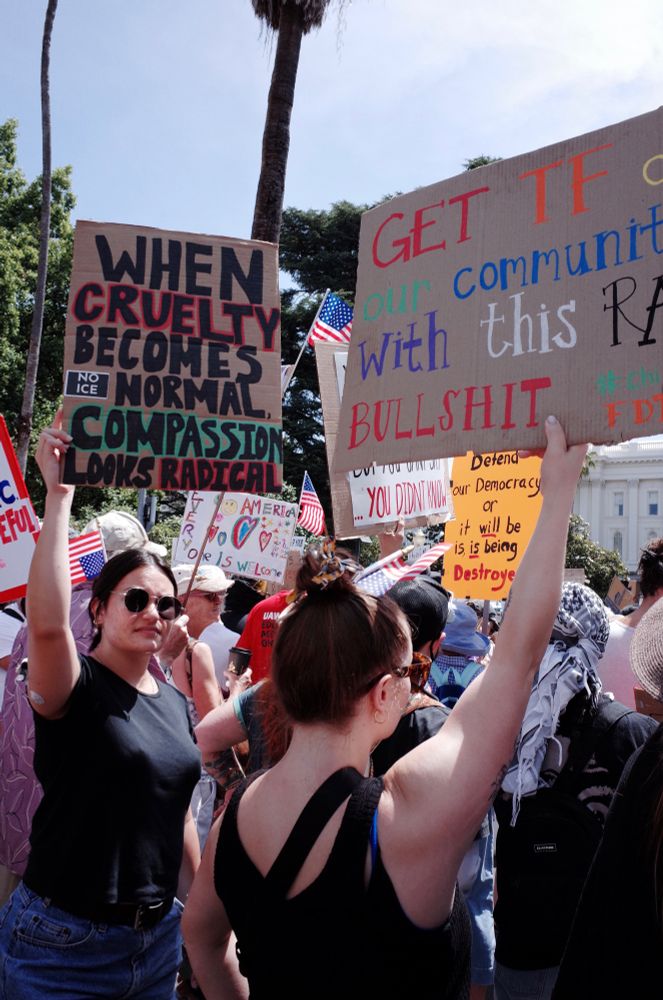 A woman in sunglasses faces the camera in a crowd of people facing away. She holds a sign that says “when cruelty becomes normal compassion looks radical” above her head. 