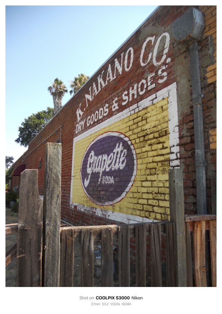 Photo of a mural on the side of an old brick building. The mural is an advertisement reading: K. NAKANO CO DRY GOODS & SHOES in block letters. Below the letters, a large yellow rectangle is painted with a purple oval in the center that reads: Grapette SODA. In the for ground an old wooden fence frames the mural with uneven wooden posts. 

 Shot on COOLPIX S3000 Nikon
 27mm f/3.2 1/320s ISO80