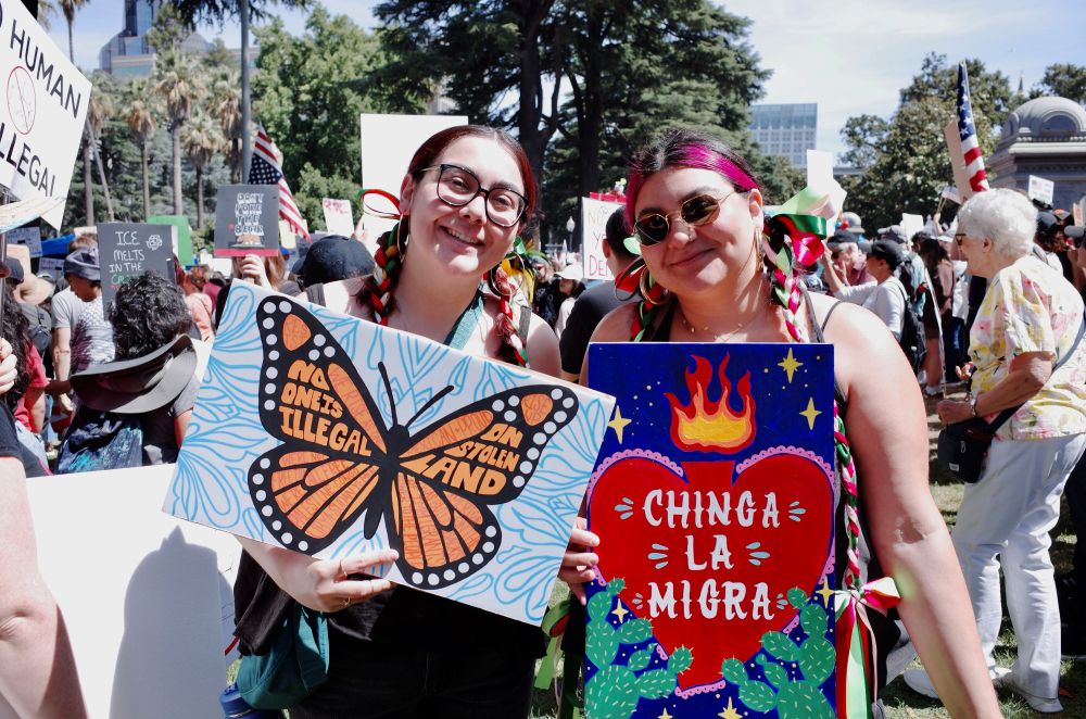 Two women smile and hold up their signs to the camera. Their hair is braided on both sides with long ribbons the colors of the Mexican flag. One holds a sign of a butterfly with the words “No one is illegal on stolen lands” painted onto its wings. The other woman’s sign is of a painted Sacred Heart symbol with the words “Chinga La Migra” painted over the heart. Nopal cacti border the heart to either side and yellow stars decorate the purple background. 