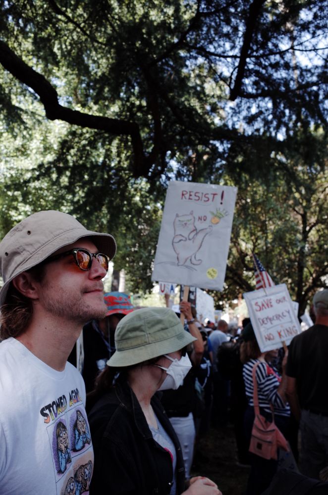A man and woman wearing bucket hats and sunglasses stand in a crowd under a large tree. A sign in the background depicts a cat karate kicking an orange with a crown. The word “Resist” is written on the top.  