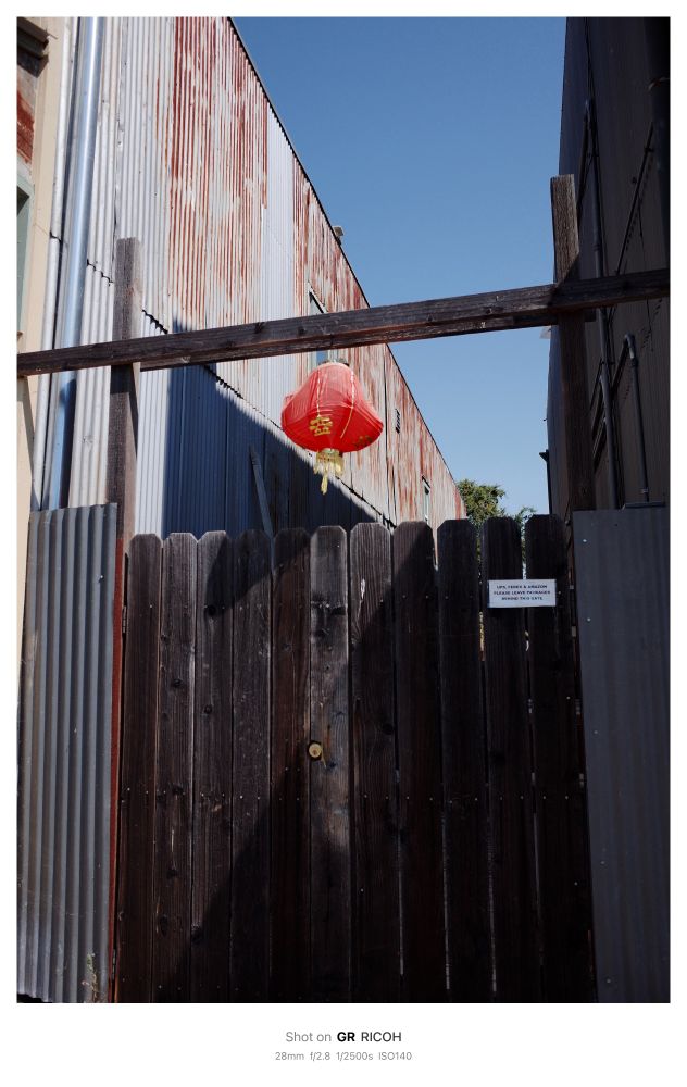 A red Chinese lantern hangs from wooden framing over a wooden gate. Behind the lantern is a large industrial building lined in rusted corrugated metal. A triangle of blue sky is visible at the top of the image.
 
Shot on GR RICOH
 28mm f/2.8 1/2500s ISO140