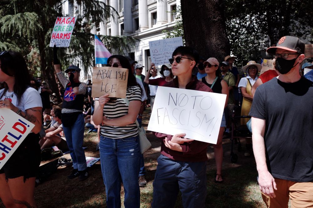 A woman hides her face behind a sign that says “Abolish ICE” next to her a man holds a sign that says “No to fascism”. A transgender pride flag is visible behind them next to another protester who holds a sign that reads “MAGA greed harms children & families”.