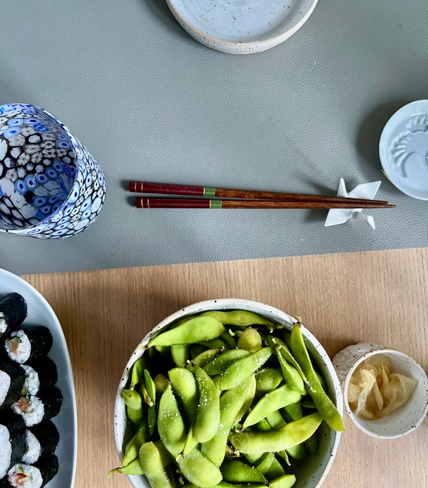 Gedeckter Tisch, Sushi, Edamame, Ingwer, ein graues platzset mit murano Glas drauf, Stäbchen auf Kranich Soja Schale und Teller 