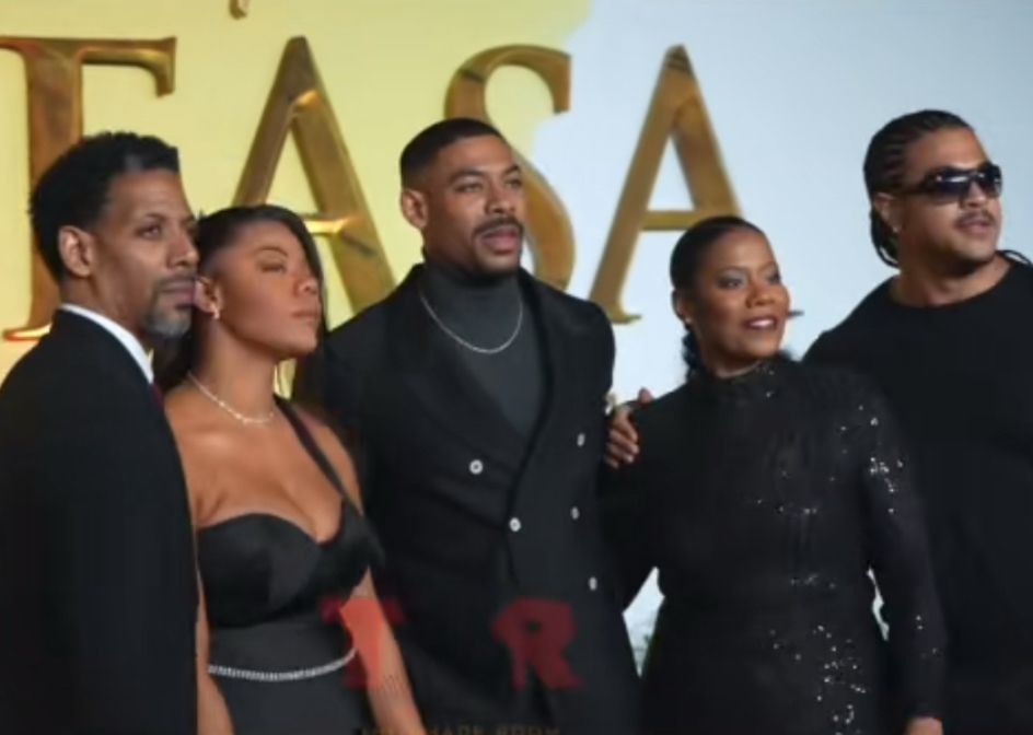 A family of ancient Black vampires in stunning black tie on the Mufasa red carpet. the photo has a shade room watermark on it, but you can ignore that. The family of vampires is beautiful. They are standing, from left to right, an elder vampire, presumably Aaron Pierre’s father, a young woman vampire, possibly Aaron pierre’s sister, a regal looking woman vampire, possible Aaron Pierre’s mother and a vampire with straight back braids and sunglasses, presumably Aaron Pierre’s brother. 