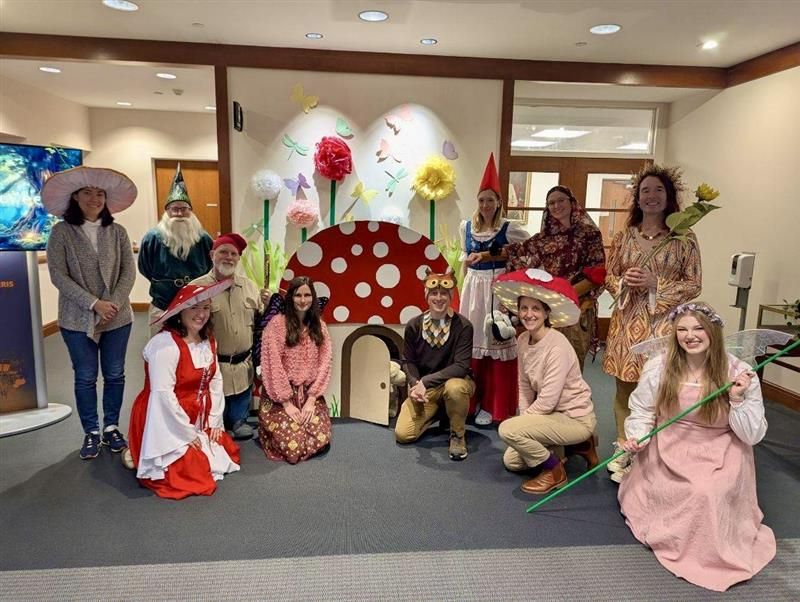 A group of people dressed as fairies, mushrooms, and gnomes posing in front of a flower backdrop and a mushroom house.
