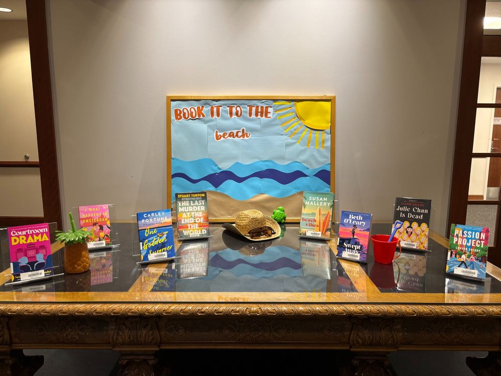 A book display on a large table featuring eight books. Signage reads, “book it to the beach.” Books featured from left to right: Courtroom Drama by Neely Tubati Alexander, What Happens in Amsterdam by Rachel Lynn Solomon, One Golden Summer by Carley Fortune, The Last Murder at the End of the World by Stuart Turton, Beach Vibes by Susan Mallery, Swept Away by Beth O’Leary, Julie Chan is Dead by Liann Zhang, and Passion Project by London Sperry.