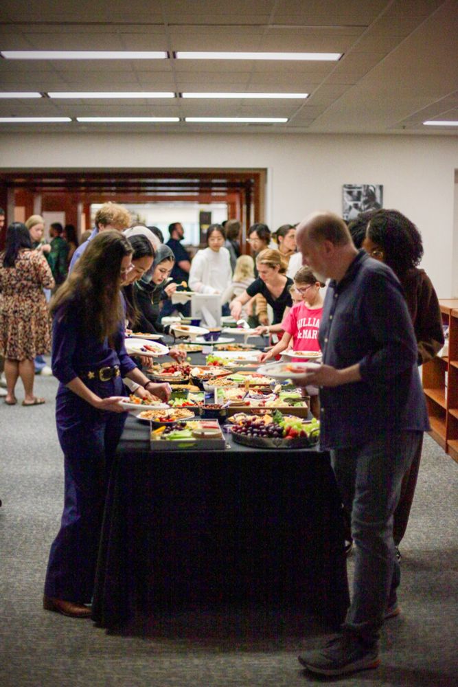 People lined on both sides of a buffet table making plates of fresh produce, cheese, and crackers.
