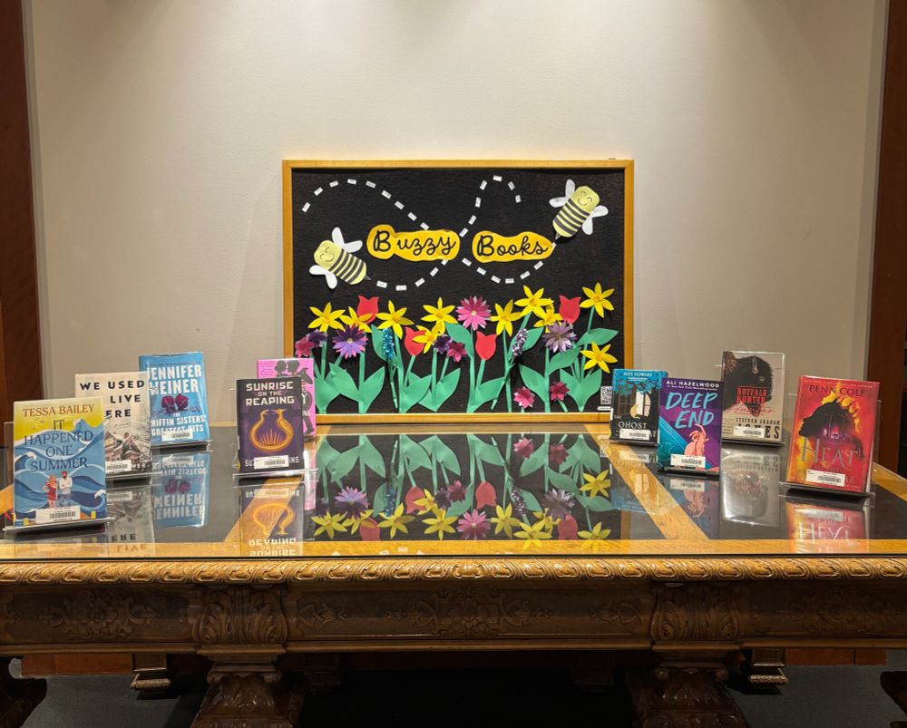 A book display featuring nine books on a large table. Signage reads, “Buzzy Books,” and features flowers and bumblebees.