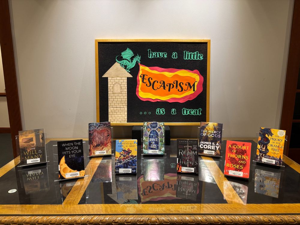 A display of 9 books on a large table. Signage features fantasy elements and says, “have a little escapism… as a treat.”