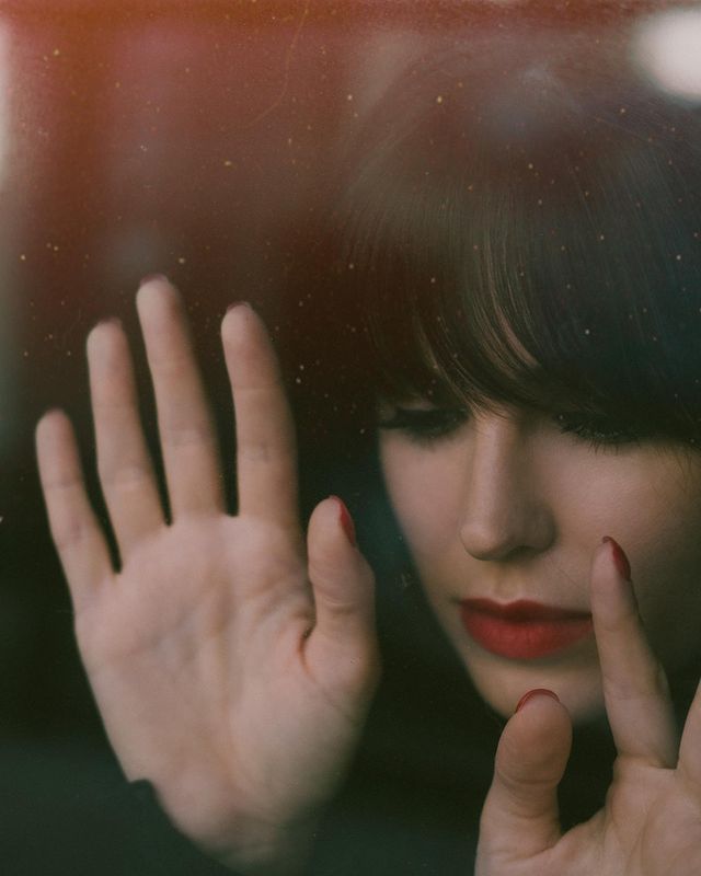 A woman with dark hair, red lipstick, nails painted red, and fair skin, with her hands on a window, looking outside.