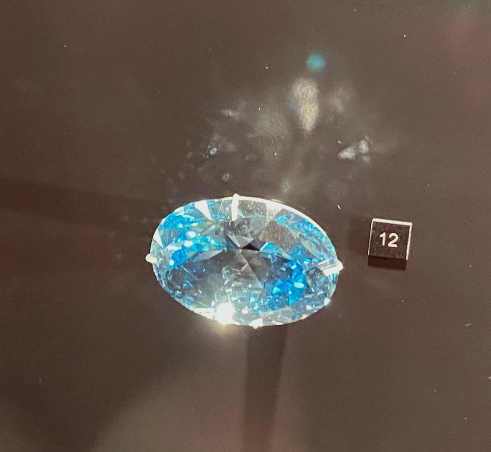 Bright blue oval cut gem (long side right to left) mounted on a black background and with a small black square label with a white 12 on it. At the Musée de Minéralogie Mines Paris.