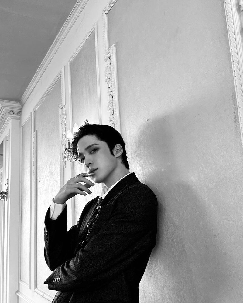 a black and white photo of jeong yunho. his head is tilted. he's still looking at the camera like he's going to fuck it into next week. this time his hand is at his mouth. his deliciously long index finger toys with his bottom lip like he's toying with my heart.