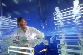 Picture of a scientist in a white lab coat in a blue room; near him are piled clear rectangles that look like glass.
