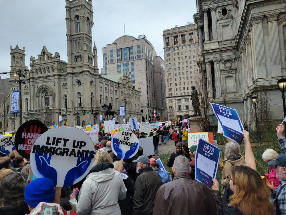 A crowd gathered by City Hall in Philadelphia. The nearest signs are round and say "lift up immigrants", "lift up choice", etc. 