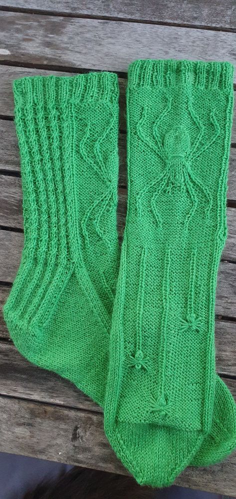 A pair of hand-knitted socks. The yarn is poisonous green. The legs has one large cabled spider, and the foot three tiny baby spiders.