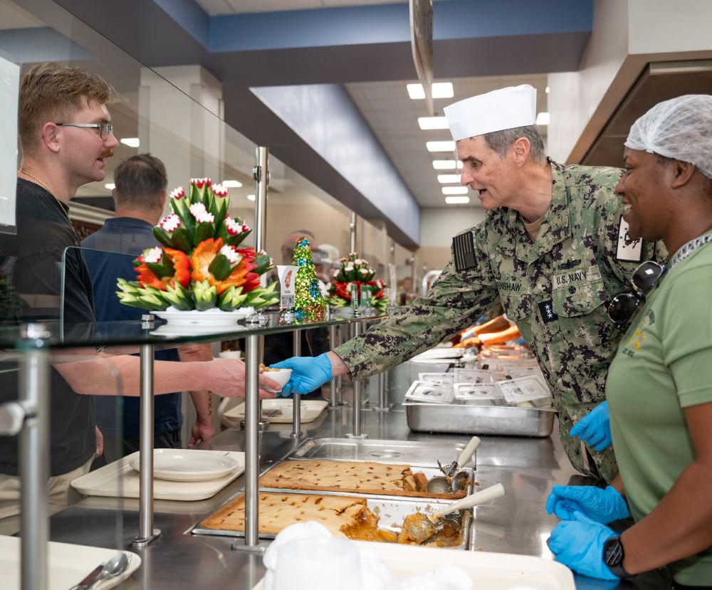 A Navy officer in camouflage uniform hands a bowl of food to a man with a tray through the window of a military dining facility's service counter.