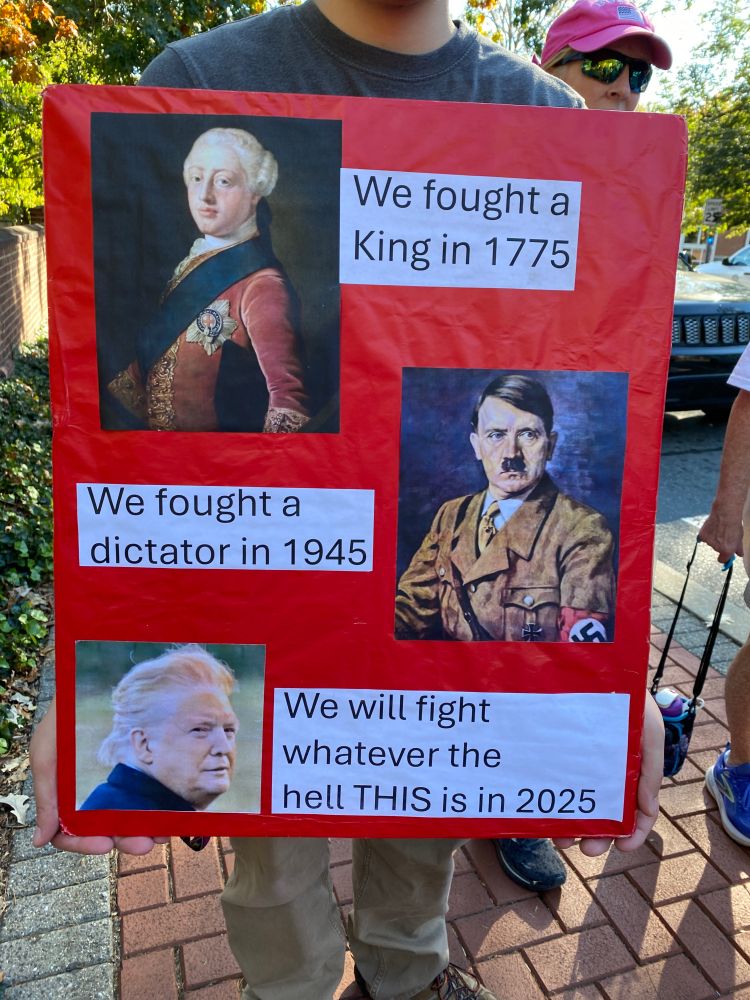 Red sign with picture of King George beside text: We fought a King in 1775; a picture of Hitler beside text: We fought a dictator in 1945; and a photo of Trump, his weird hair blown back to reveal even weirder makeup line, beside text: We will fight whatever the hell THIS is in 2025.