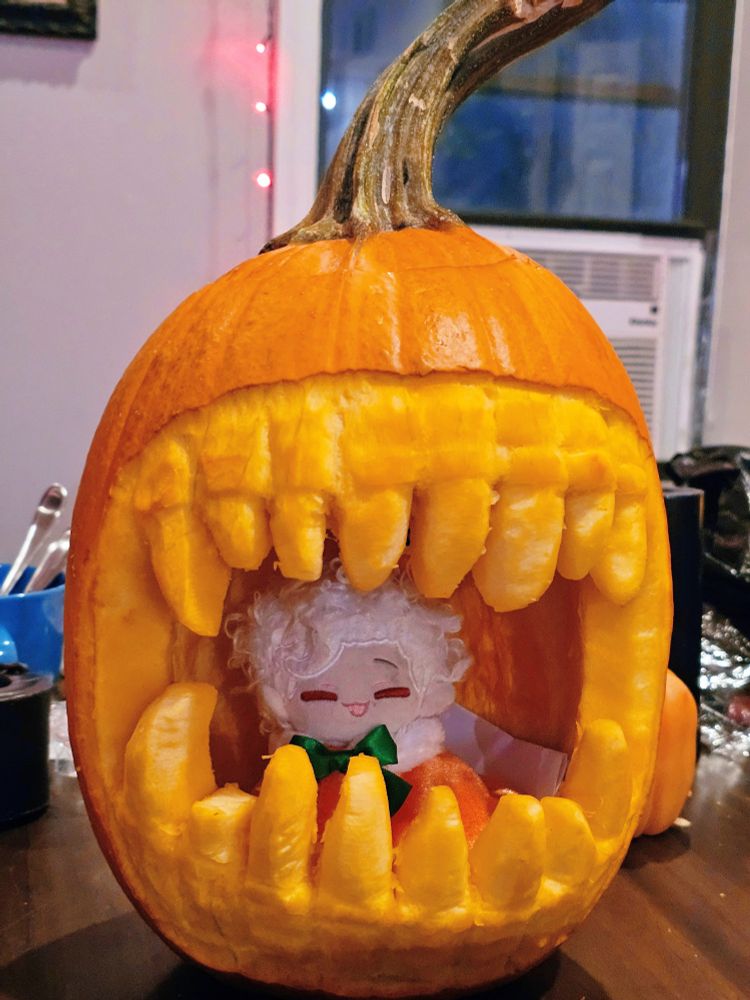 photo of a giant mouth with sharp teeth carved into a pumpkin. a smiling astarion plushie in a pumpkin outfit is inside it.