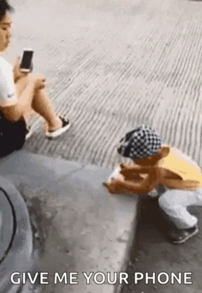 a woman is sitting on the sidewalk next to a child who is kneeling down and playing a game on his phone .