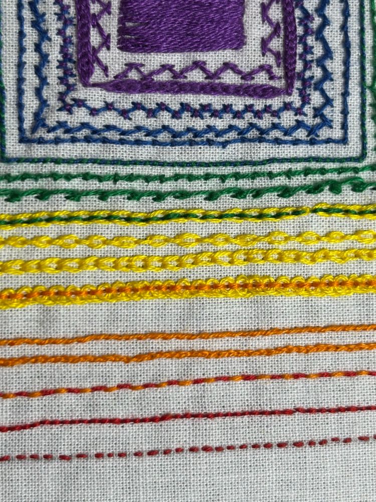 A close up photo of the sampler showing the individual lines of stitches. From bottom to top: Running Stitch and Backstitch in red, Holbein Stitch in red and orange, Split Stitch and Outline Stitch in orange, Pekinese Stitch in orange and yellow, Chain Stitch and Cable Chain Stitch in yellow, Backstitched Chain Stitch in yellow and green, Scroll Stitch & Pearl Stitch in green, Whipped Backstitch in green and blue, Coral Stitch and Herringbone Stitch in blue, Tied Herringbone Stitch in blue and purple, and Hungarian Braided Chain Stitch, Chevron Stitch, and Satin Stitch in purple.