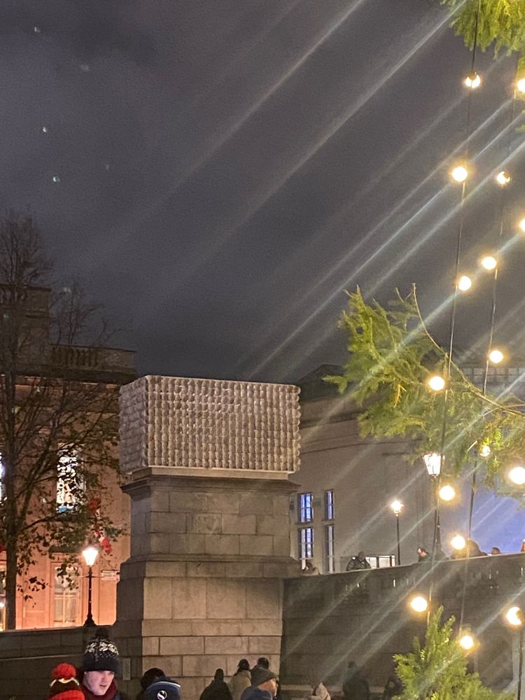 The fourth plinth in Trafalgar Square with diagonal lines of light reflecting from the Christmas tree from Oslo. Mil Veces un Instante is there name of the art work on the plinth and is made up of plaster casts of the faces of 726 trans, non-binary, and gender non-conforming people.