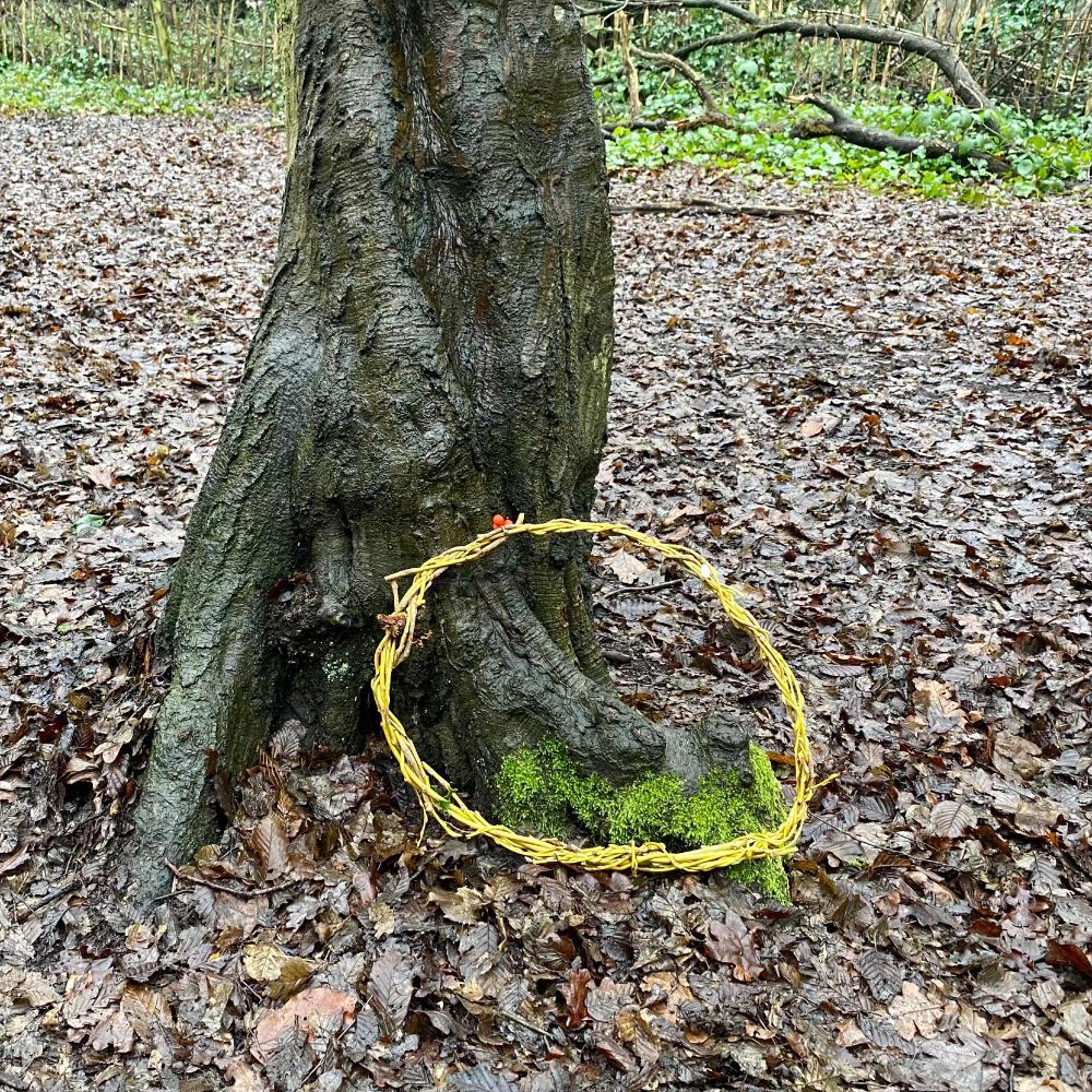 Woven willow circle placed by a moss covered tree trunk. Created during a circle of trees environmental art group at Highgate’s woods. The ground is wet with rain and fallen leaves, we found the flow of water and gathered ourselves, from the mountain streams to the rivers and bogs to the lakes and shore of the sea. The might oceans, mist, clouds, rain and circling round to streams.  The willow has holly, berries, bark and a snow drop 