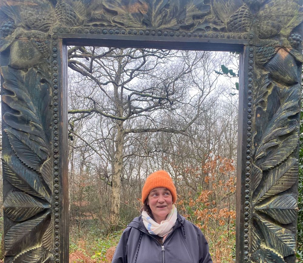 Me standing in the centre of the wooden frame pictures in previous image a silver birch in the distance and green and orange leaves around. I am practicing self care keeping warm and wondering 