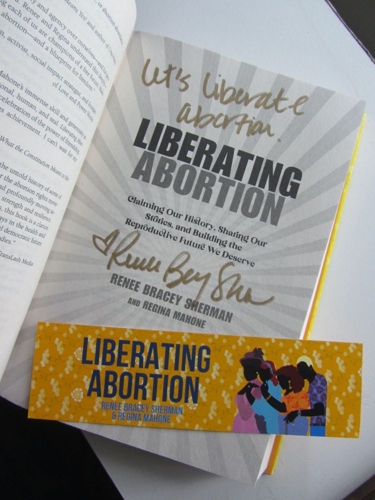 The inside title page of the book "Liberating Abortion: Claiming Our History, Sharing Our Stories, and Building the Reproductive Future We Deserve Hardcover" by Renee Bracey Sherman and Regina Mahone. The title page is signed by Renee Bracey Sherman and she also wrote "Let's liberate abortion." There is an orange "Liberating Abortion" bookmark lying on the bottom of the title page. The bookmark has the figures of four Black people from different time periods on it.