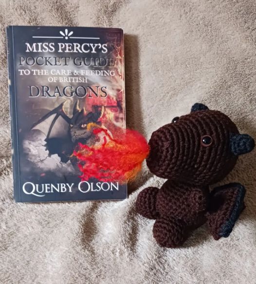 A brown and black crocheted dragon breathing (yarn) fire in front of a copy of the book Miss Percy's Pocket Guide. 
