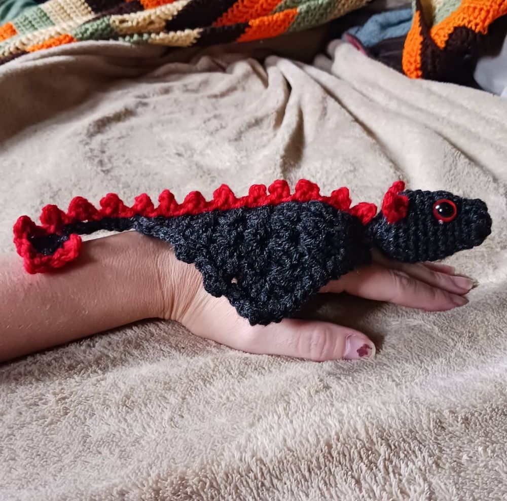 A crocheted black dragon with red horns, eyes, and spikes, resting on the back of someone's hand. 
