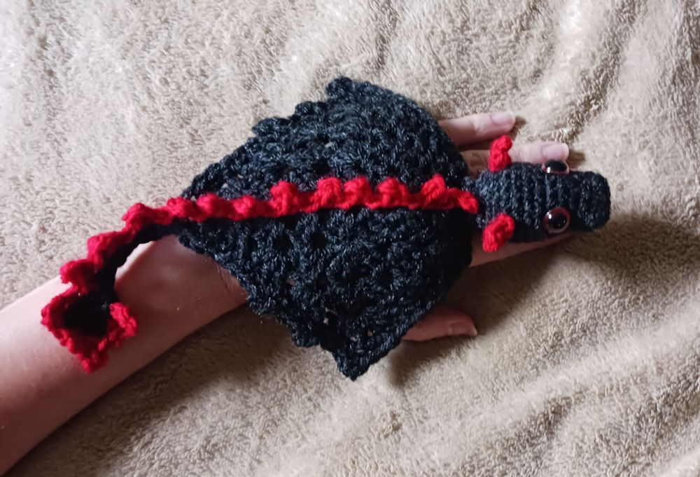 A crocheted black dragon with red horns and spikes, resting on the back of someone's hand. 