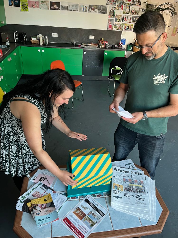 A colour photograph of two people standing at a low coffee table in a community space. On the table a green tombola decorated with yellow diagonal stripes. On the left a young white woman in her 20s with long black hair wearing a sleeveless black and white floral dress leans over the tombola about to reach inside it. On the right a tall white man in his 40s wearing a dark green T-shirt and spectacles holds a small piece of paper. He has just drawn out of the tombola.