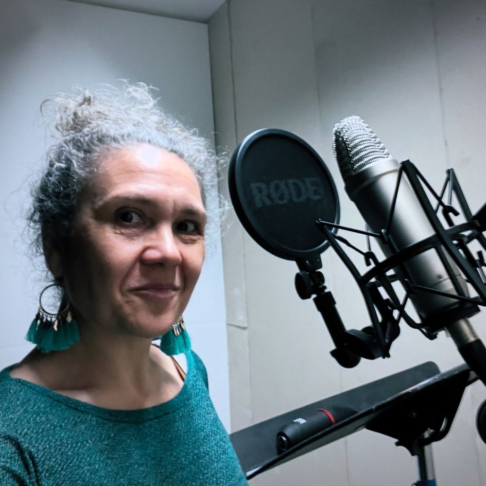 Colour photo of a white woman in her 40s with dark curly hair tied up on top of her head, and dark eyes. She standing in a white soundproof booth by a silver microphone on a stand behind a black circular pop-shield. She wears a green jumper and feathery green dangly earrings and is smiling. 