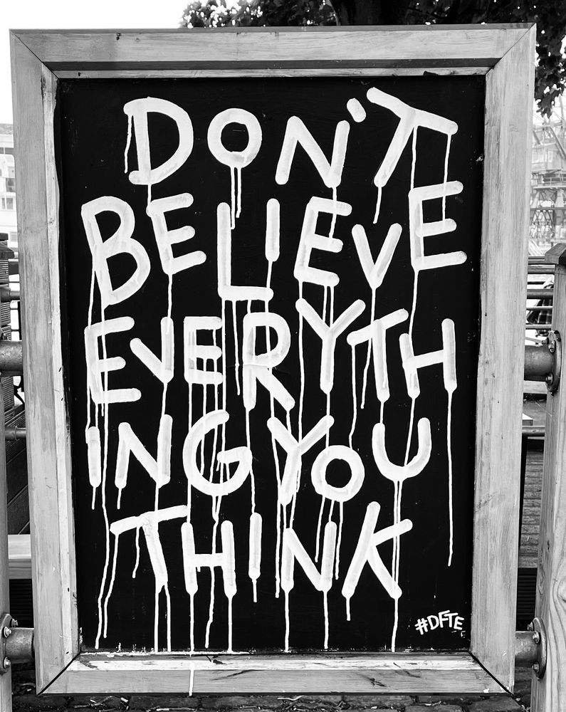 A black and white photograph of a large portrait orientated blackboard. Written in white the words don’t believe everything you think. The white paint used for the letters runs and leaves little vertical trails on the blackboard. The blackboard is fixed between two wooden fence posts.