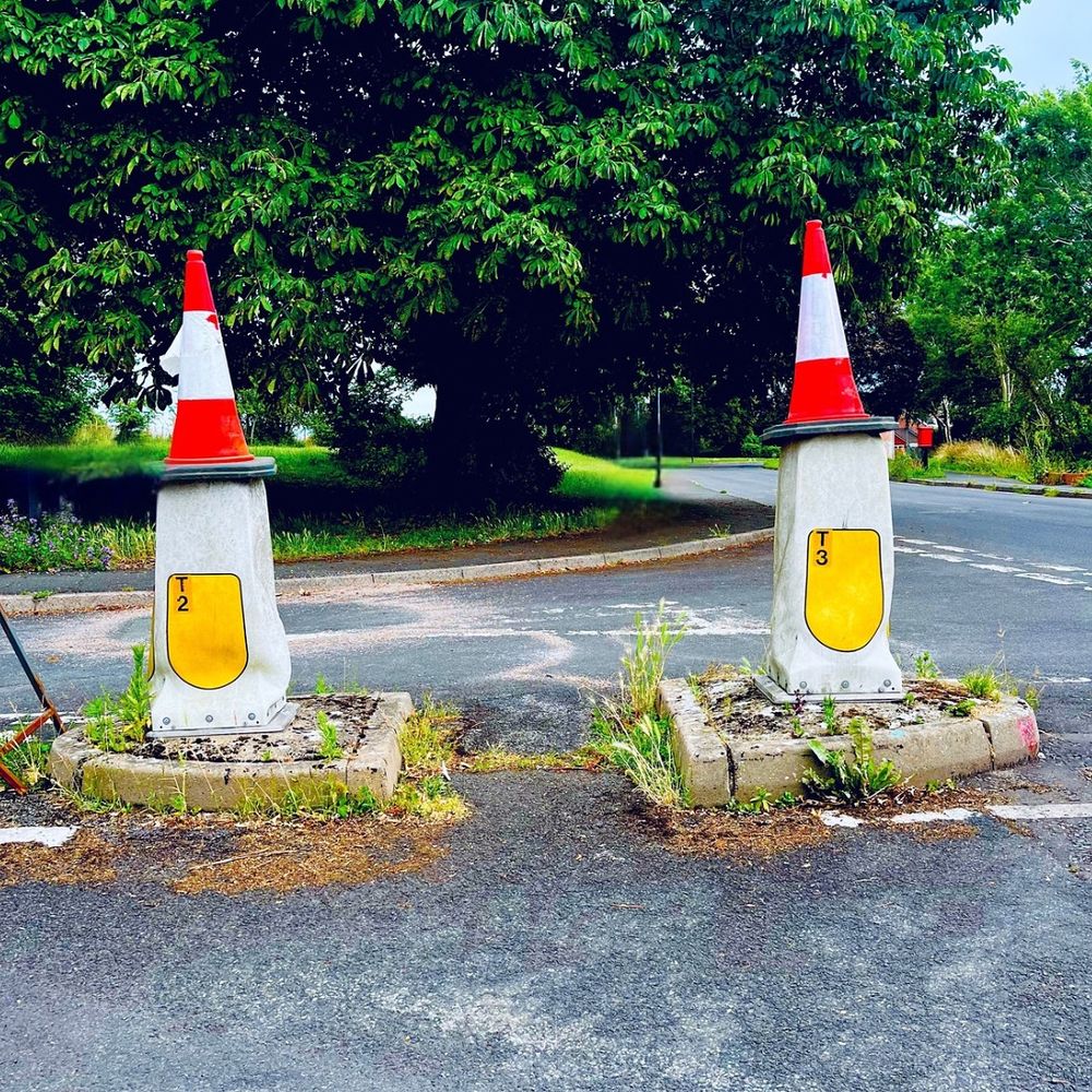 Colour photo of two square white plastic bollards on a road junction. Each bollard has a red traffic cone balanced on top. Green trees beyond.