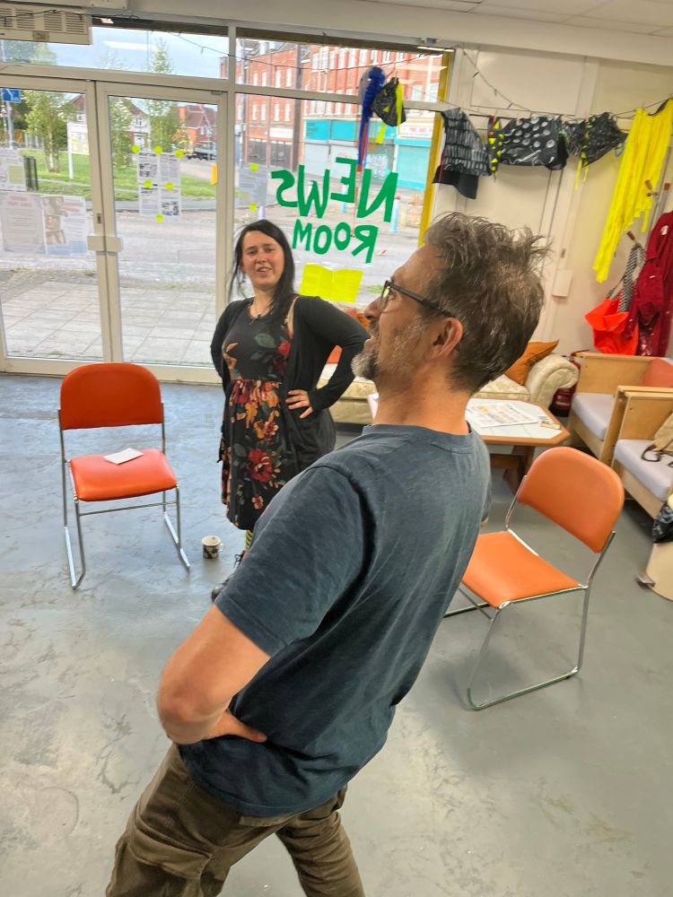 A colour photograph of two people in a community space doing a theatre improv exercise. In the foreground a tall white man in his 40s with a dark green T-shirt and short dark hair and glasses stands with his hands on his hips, leaning backwards with a proud expression. Beyond a white woman in her 20s with long black hair, wearing a long sleeved black top and a black skirt with red floral print mimics the man’s pose.