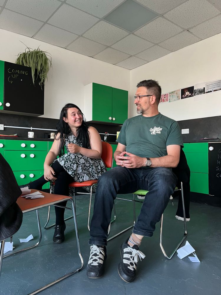 A colour photograph of two people in a community space in the act of a theatre improv exercise. They are both sitting. On the left a young white woman in her 20s with long black hair wearing a sleeveless black and white floral dress smiles at the man sitting next to her. On the right a tall white man in his 40s wearing jeans, a dark green T-shirt and spectacles mines holding an ice cream cone and looks at the woman with a smile. In front of them another chair covered with small slips of paper. Some other slips of paper are scattered on the floor at their feet.