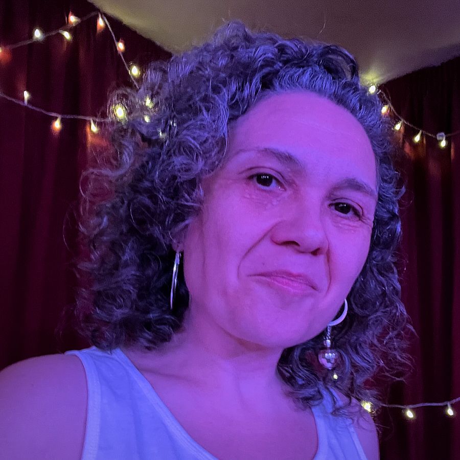 A colour head and shoulders selfie of a white woman in her 40s. She has dark curly shoulder length hair dark eyes was a white vest top and large silver hoop earrings. She is smiling. Behind her red theatre curtains and golden fairy lights.