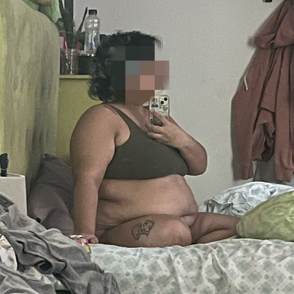 picture of me (tan, fat, dark brown hair with faded teal tips) sitting in bed criss cross. i’m wearing tan underwear that’s not really visible and a green cotton bra. my face is pixelated 