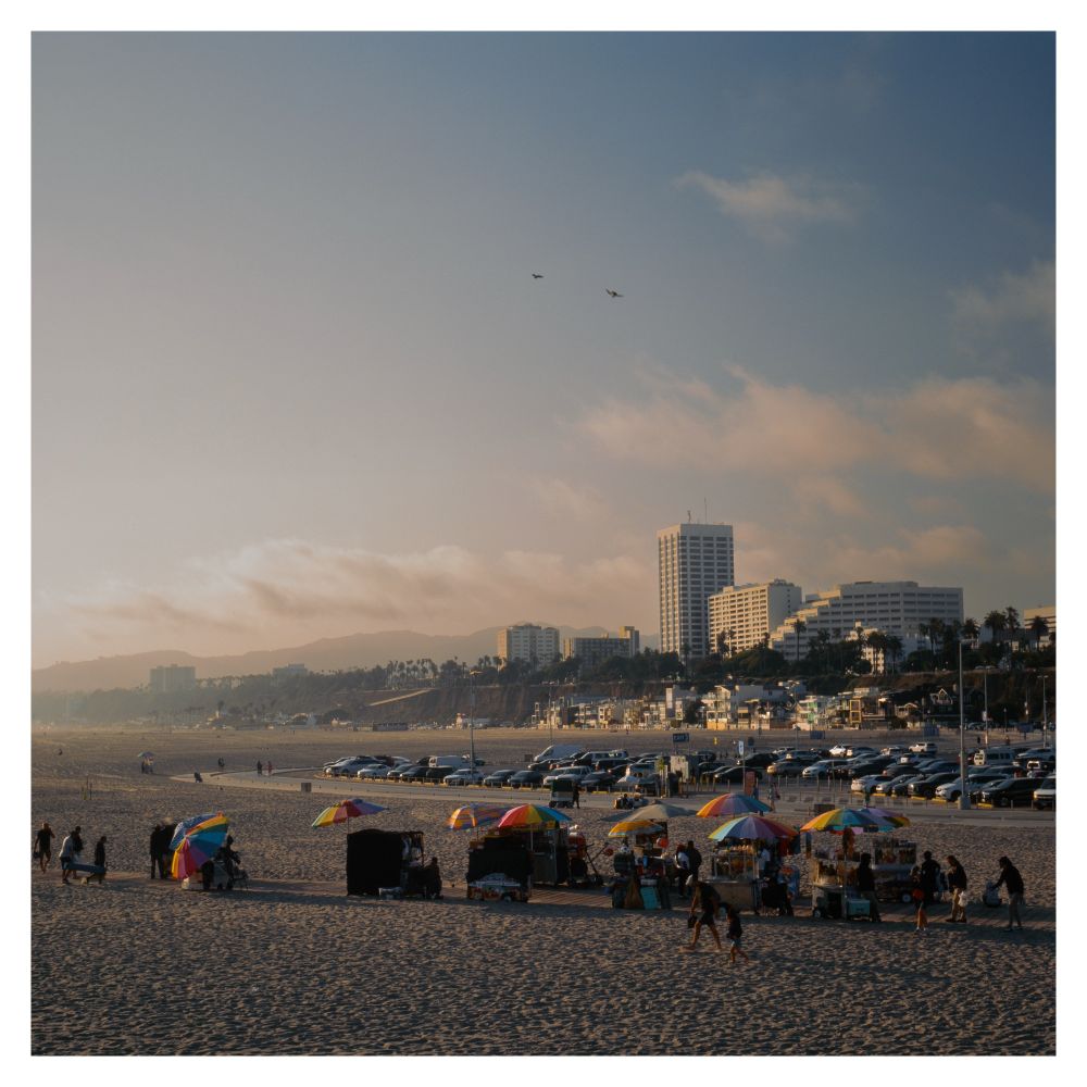 A row of street vendors with rainbow sun umbrellas on the Santa Monica beach. In the background are buildings in a row and lines of cars parked by beachgoers. 