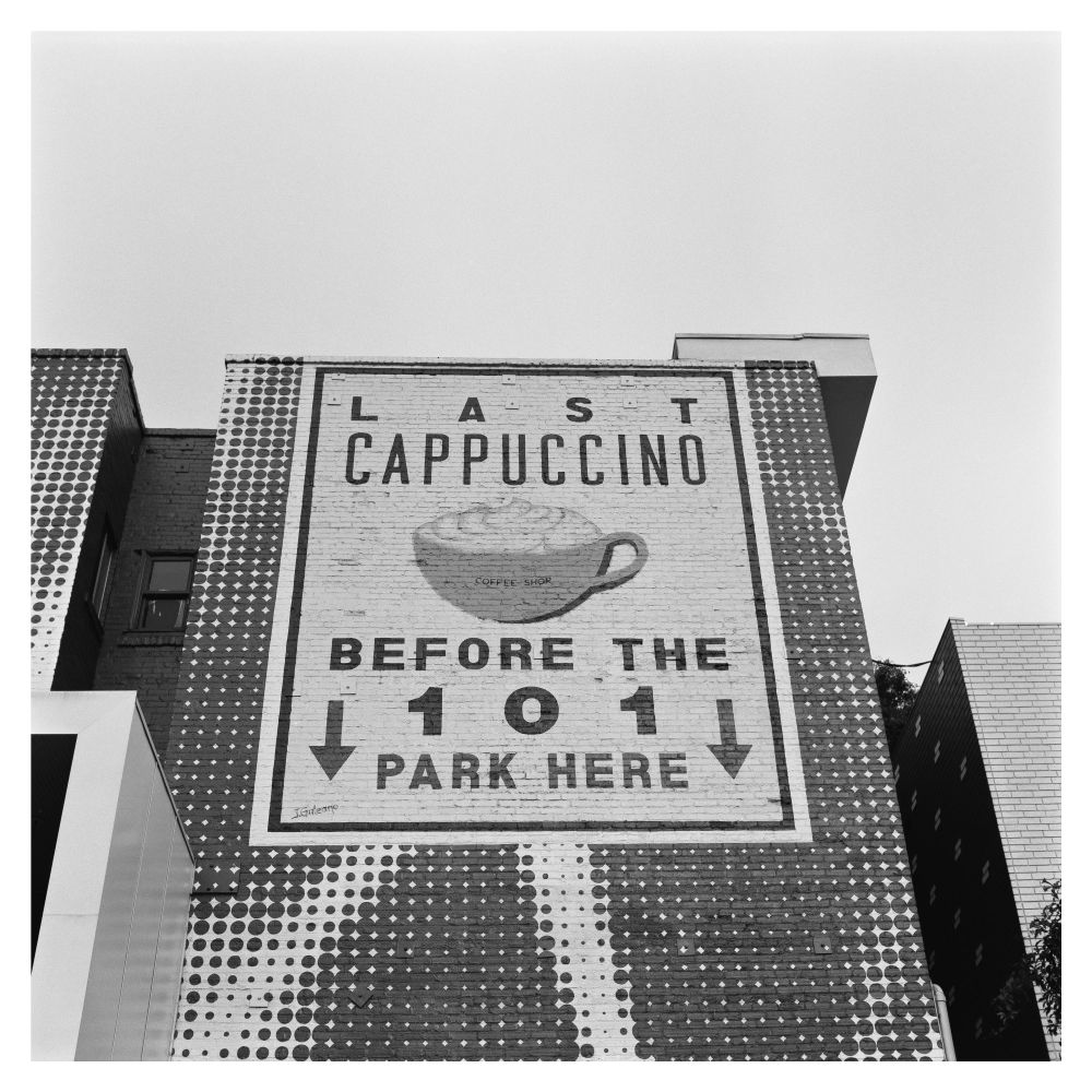 A sign painted on the Hotel Adler/Franklin with an image of a cappuccino with the words “LAST CAPPUCCINO BEFORE THE 101 PARK HERE”. 