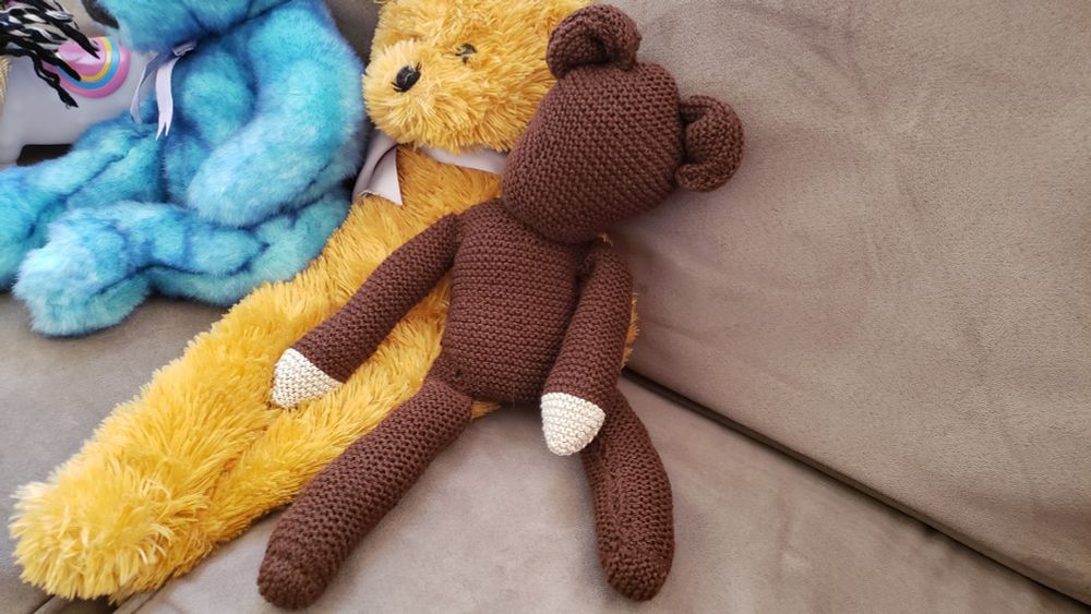 3 stuffed bears sitting on a couch. Top left blue bear, middle a tan bear, and the last bear a dark brown teddy that looks like Mr Bean's Teddy without a face. 