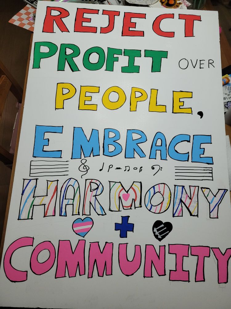 Photo of a protest sign crafted out of a white poster board. The words read "Reject Profit over People, Embrace Harmony + Community" in a variety of colors. The word "harmony" is colored in rainbow lines that ripple away from a pink heart in the center, above which are music notation symbols. Two hearts colored with the transgender flag and the anti fascist symbol of 3 downward striking arrows are arranged on either side of the "plus" symbol..