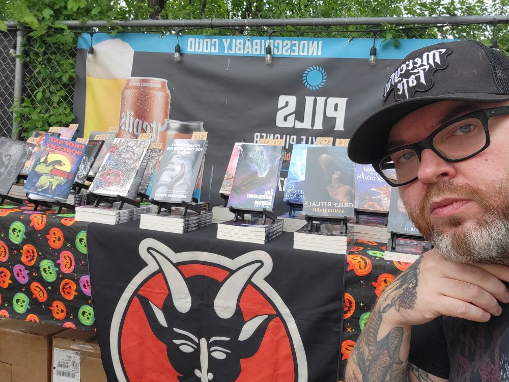 A photo of me, a grumpy old punk in a Mercyful Fate hat, in front of my table at the Utepils Pride event. The table is covered in Weirdpunk titles.