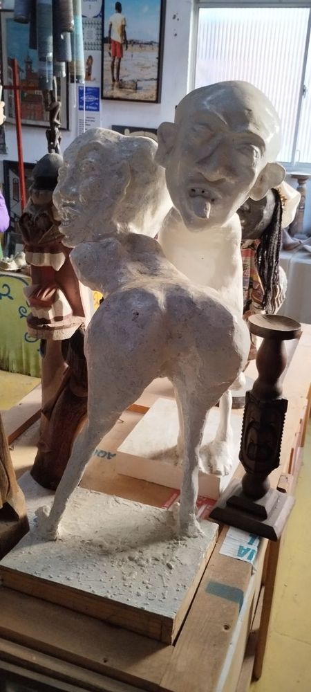 Duas esculturas brancas, uma é de uma mulher sem braços que está com o tronco abaixado e a bunda virada para a câmera. Atrás dela e de frente para a câmera, vemos um homem mostrando a língua. O rosto das duas esculturas parecem carrancas, e há uma carranca de madeira ao lado esquerdo da foto, atrás das esculturas brancas